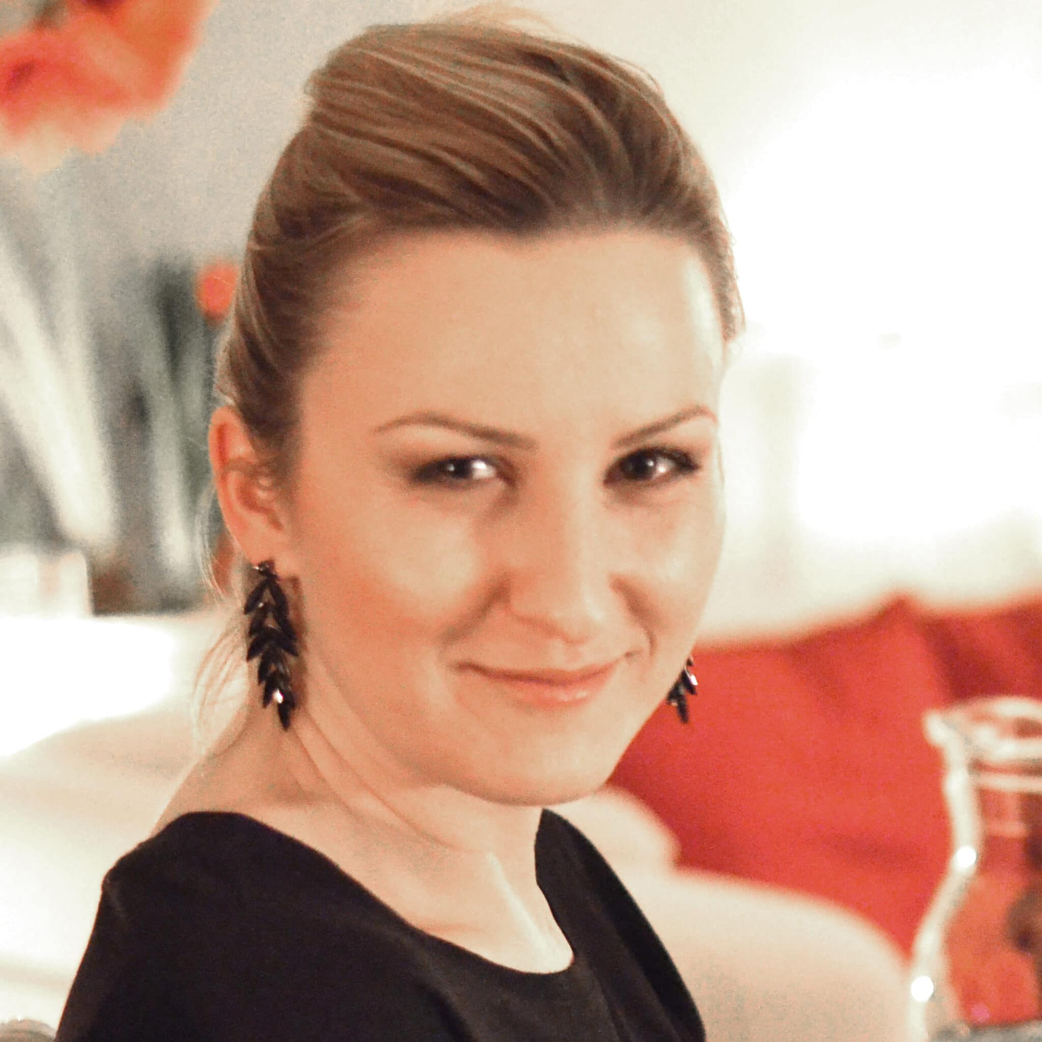 En person med en tillbakadragen frisyr, klädd i en svart topp och örhängen, ler lätt när han sitter inomhus.