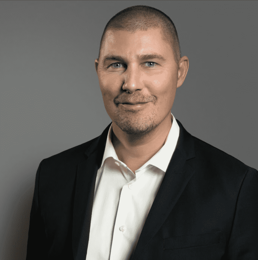 En man med kort brunt hår och ljusa ögon, klädd i vit skjorta och mörk kavaj, ler lätt mot en grå bakgrund. winwinweb