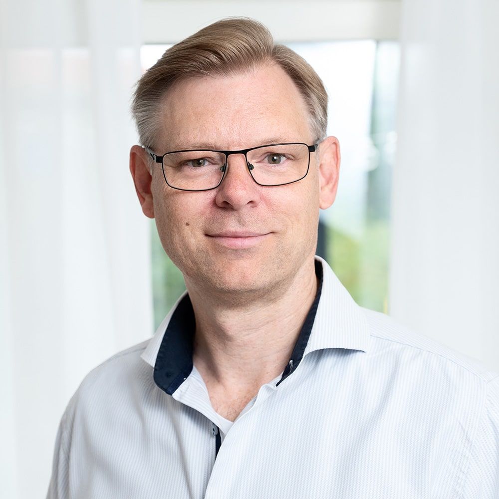 En man med kort, ljust hår och glasögon ler vänligt. Han är klädd i en ljus skjorta med krage och mörkt innerfoder. Den mjukt upplysta bakgrunden, med vita gardiner och ett fönster, ger en inbjudande ton åt vår sida om oss. winwinweb