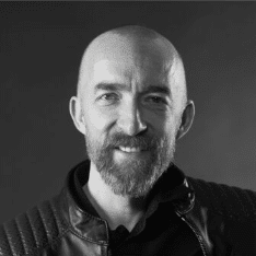 En man med skalligt huvud och helskägg ler mot kameran. Han bär en mörk, strukturerad jacka och bakgrunden är enkel och mörk, vilket skapar en professionell känsla av ett studioporträtt. winwinweb